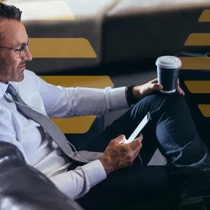 Mature businessman using a mobile phone while sitting in an airport cafe, waiting for his flight reading text message on his cell phone at first call lounge. This conveys the concept of increased passenger experience delivered by Airport Hive and Appssenger.